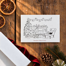 Load image into Gallery viewer, A square white greeting card with a black line drawing of a cozy holiday living room scene, placed on a rustic wooden surface with dried orange slices, a red ribbon, pine branches, and a pinecone around it. The card reads 'Season's Greetings'.
