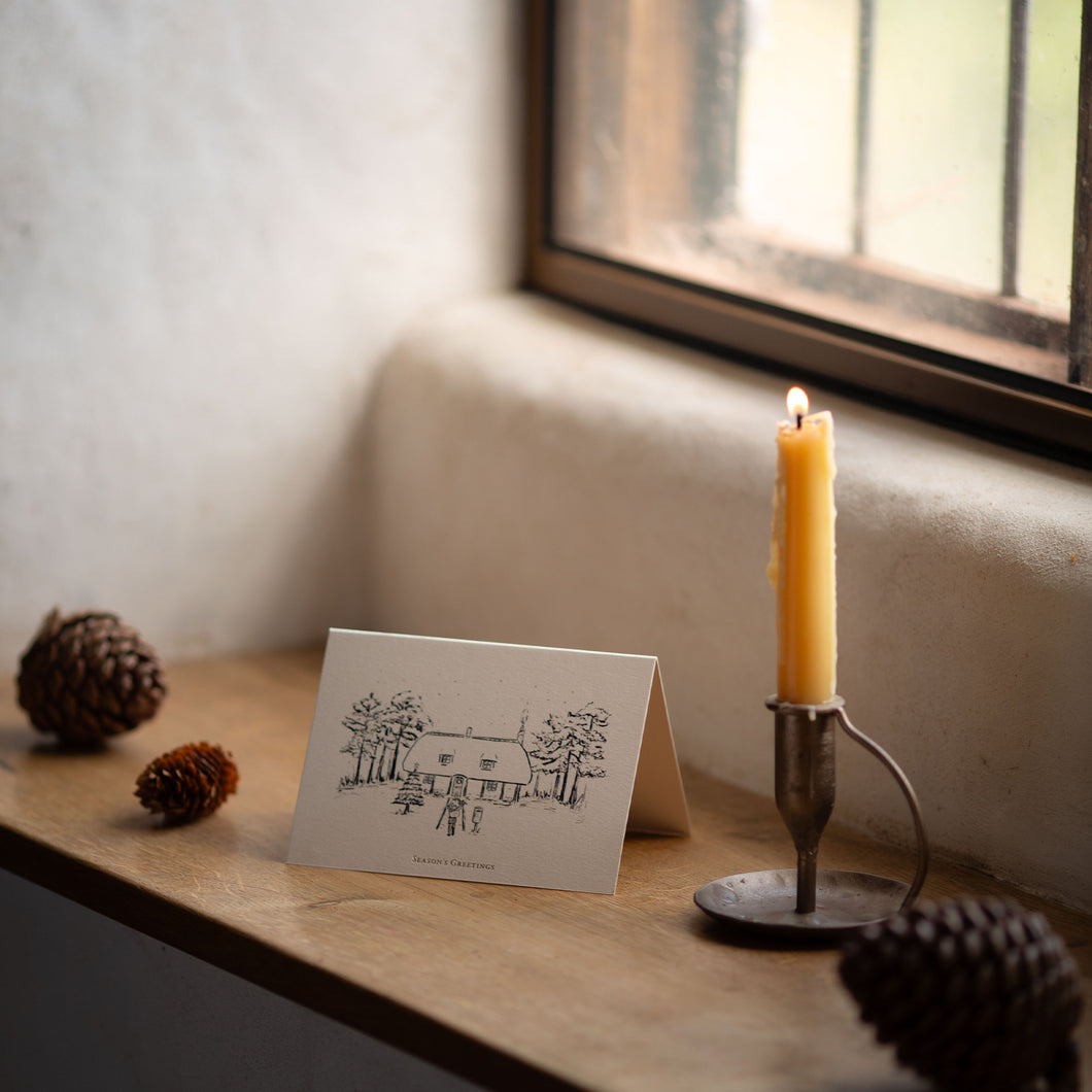 Candle in a holder next to a greeting card on a wooden surface with pinecones.
