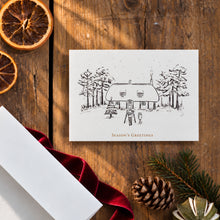 Load image into Gallery viewer, A white Christmas greeting card featuring a black line art illustration of a cottage surrounded by trees, with 'Season's Greetings' printed below. The card is styled in a flat-lay on a rustic wooden table alongside dried orange slices, a red velvet ribbon, a pinecone, and evergreen branches.
