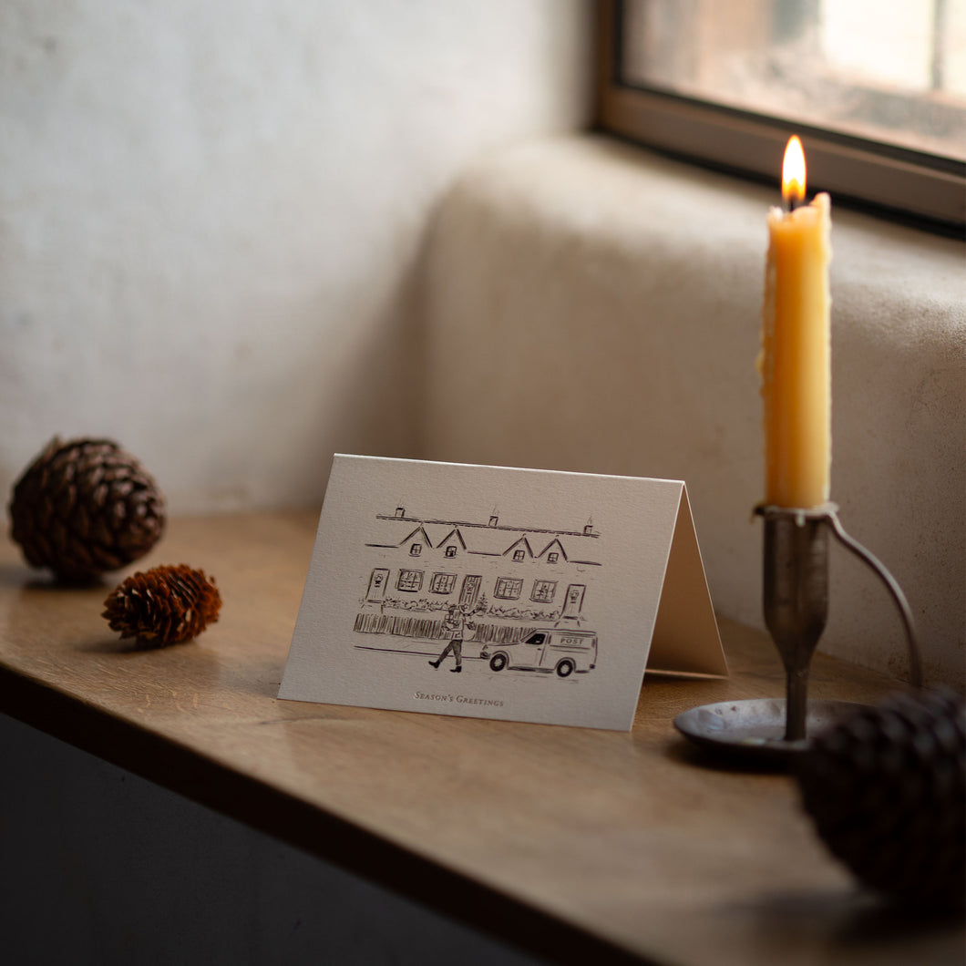 Christmas card with house illustration next to a lit candle on a wooden surface.