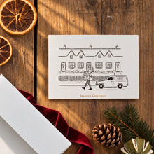 Load image into Gallery viewer, A holiday greeting card with a black line drawing of a postman delivering a Christmas tree in front of row houses, styled on a rustic wooden table with dried orange slices, a red ribbon, pinecones, and evergreen branches.
